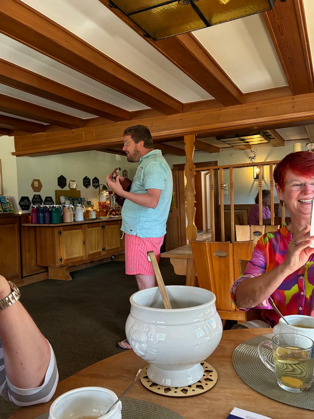 Im Speiseraum spielt ein Mann Ukulele, vorn lacht eine Gästin, auf dem Tisch steht eine weiße Suppenterrine