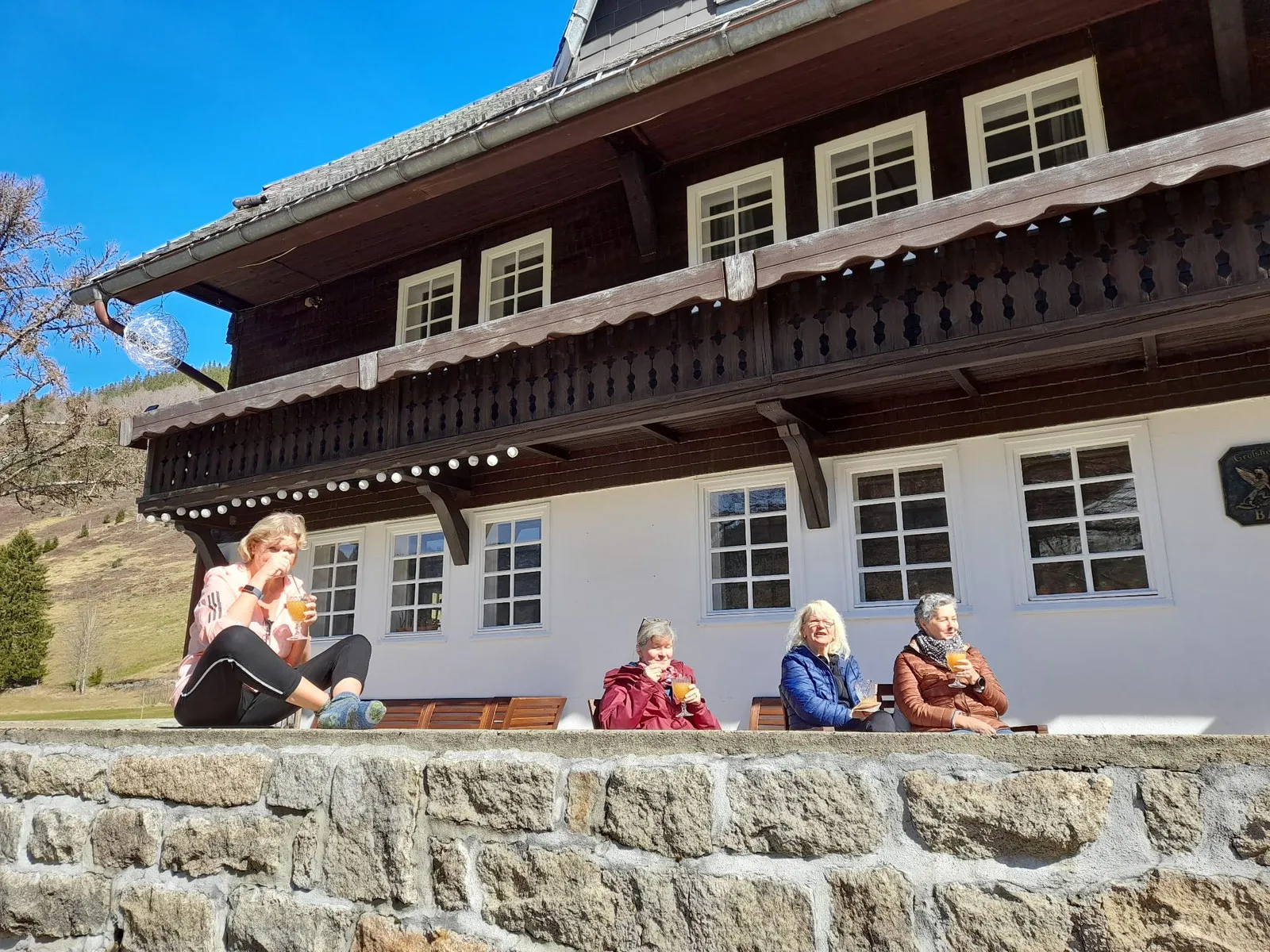 Gäste sitzen in der Sonne auf der Mauer vor der Heimatschmiede, einem großen Schwarzwaldhaus mit dunklem Holzbalkon