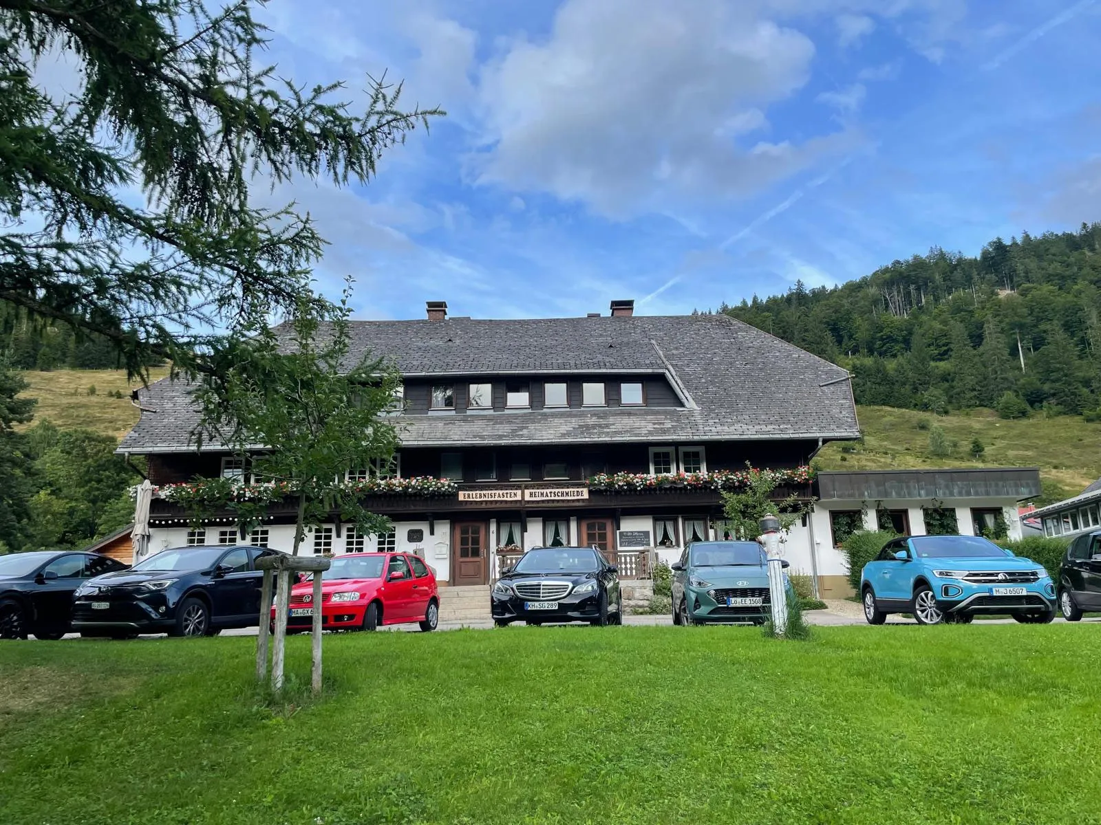 Die Heimatschmiede hinter einer Wiese, davor parken Autos auf dem Parkplatz am Haus
