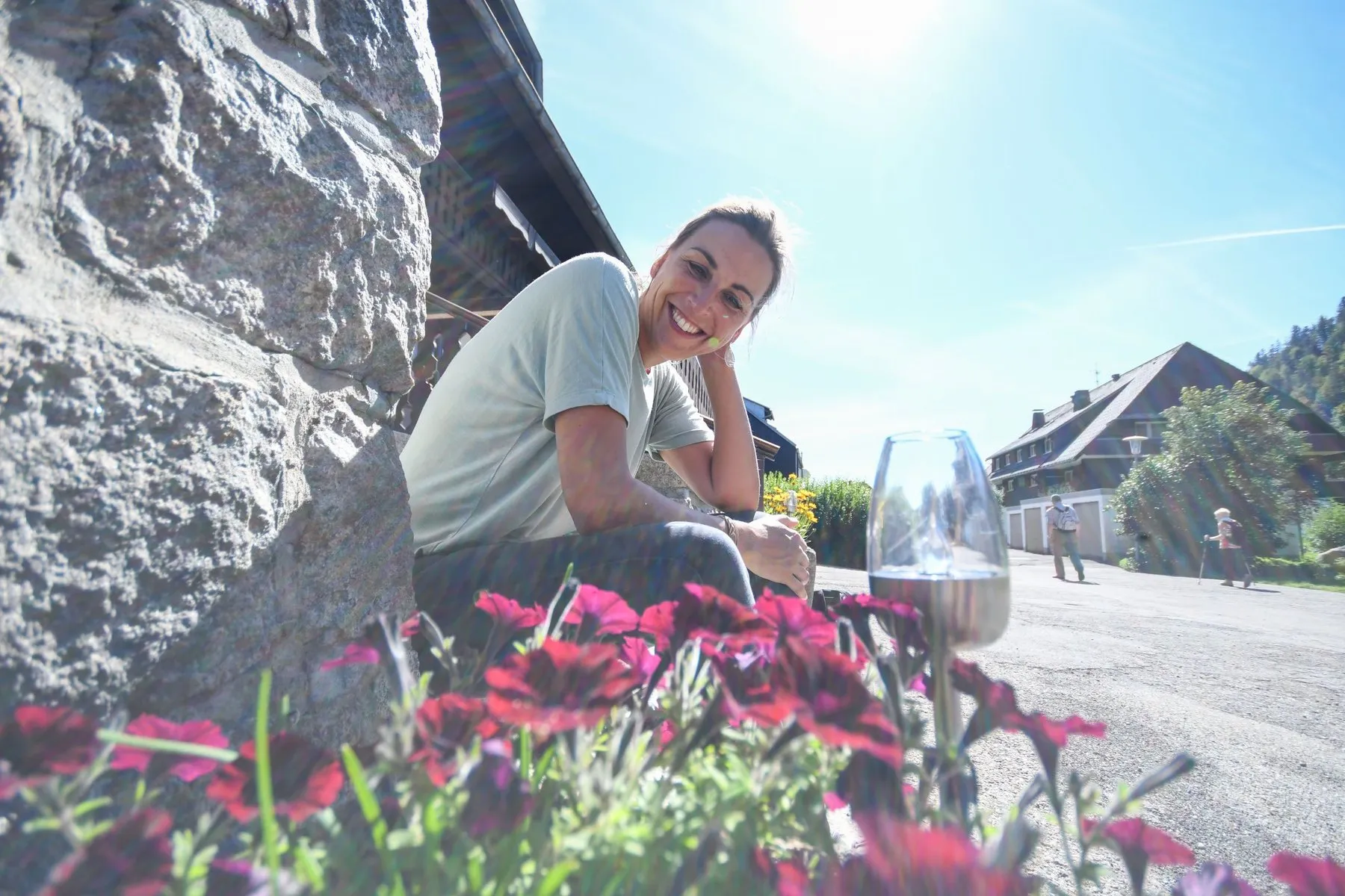Nicole Fürderer sitzt lächelnd an einer Steinmauer in Menzenschwand, vorne blühen rote Blumen, hinten ein Schwarzwaldhaus