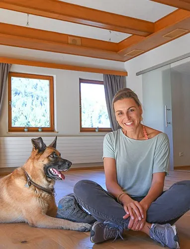 Nicole Fürderer sitzt lächelnd im Schneidersitz auf dem Holzboden, neben ihr liegt ein großer Hund