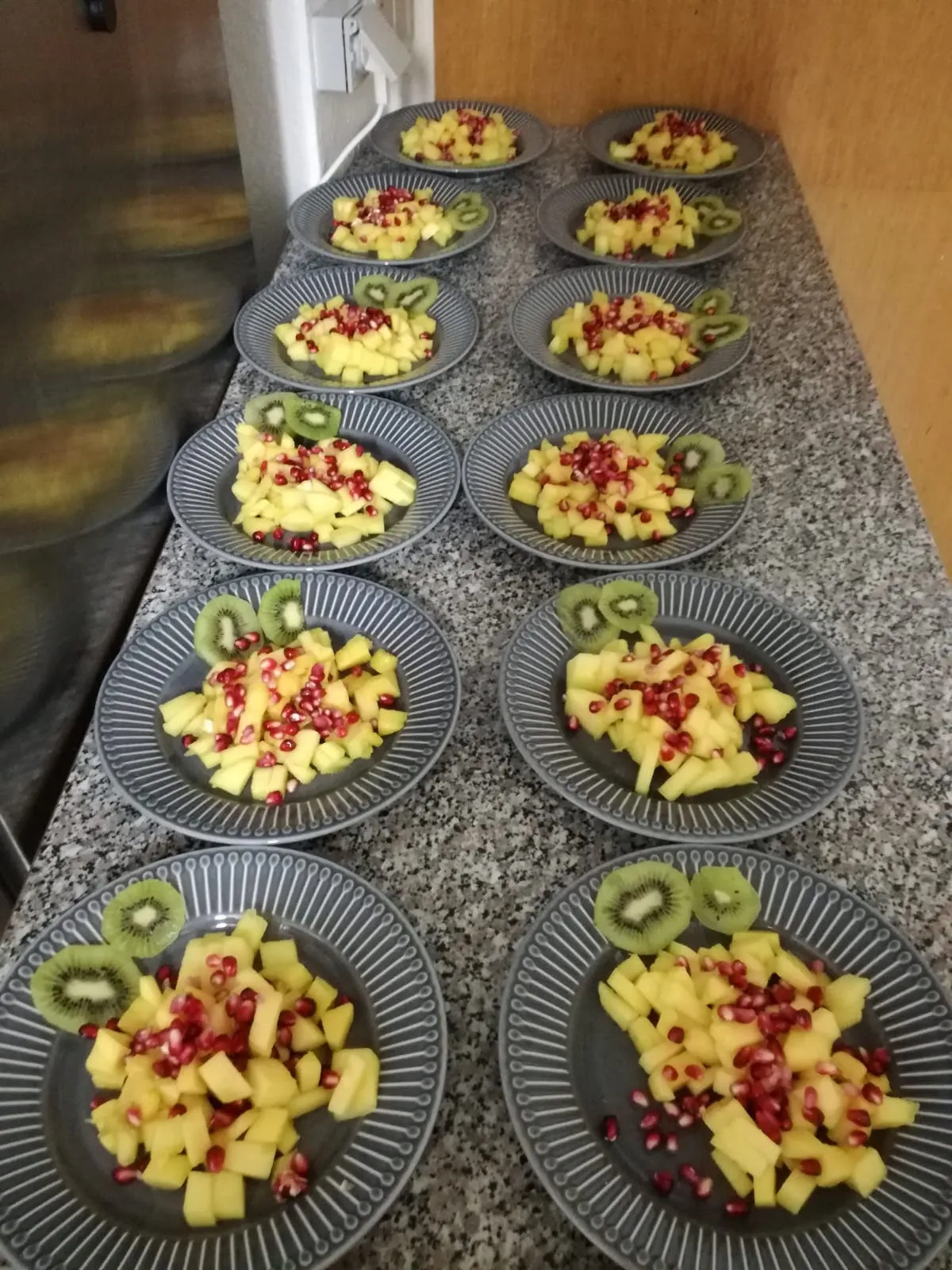 Zwölf Teller mit Obstsalat aus Mango, Kiwi und Granatapfel stehen aufgereiht auf der Küchentheke
