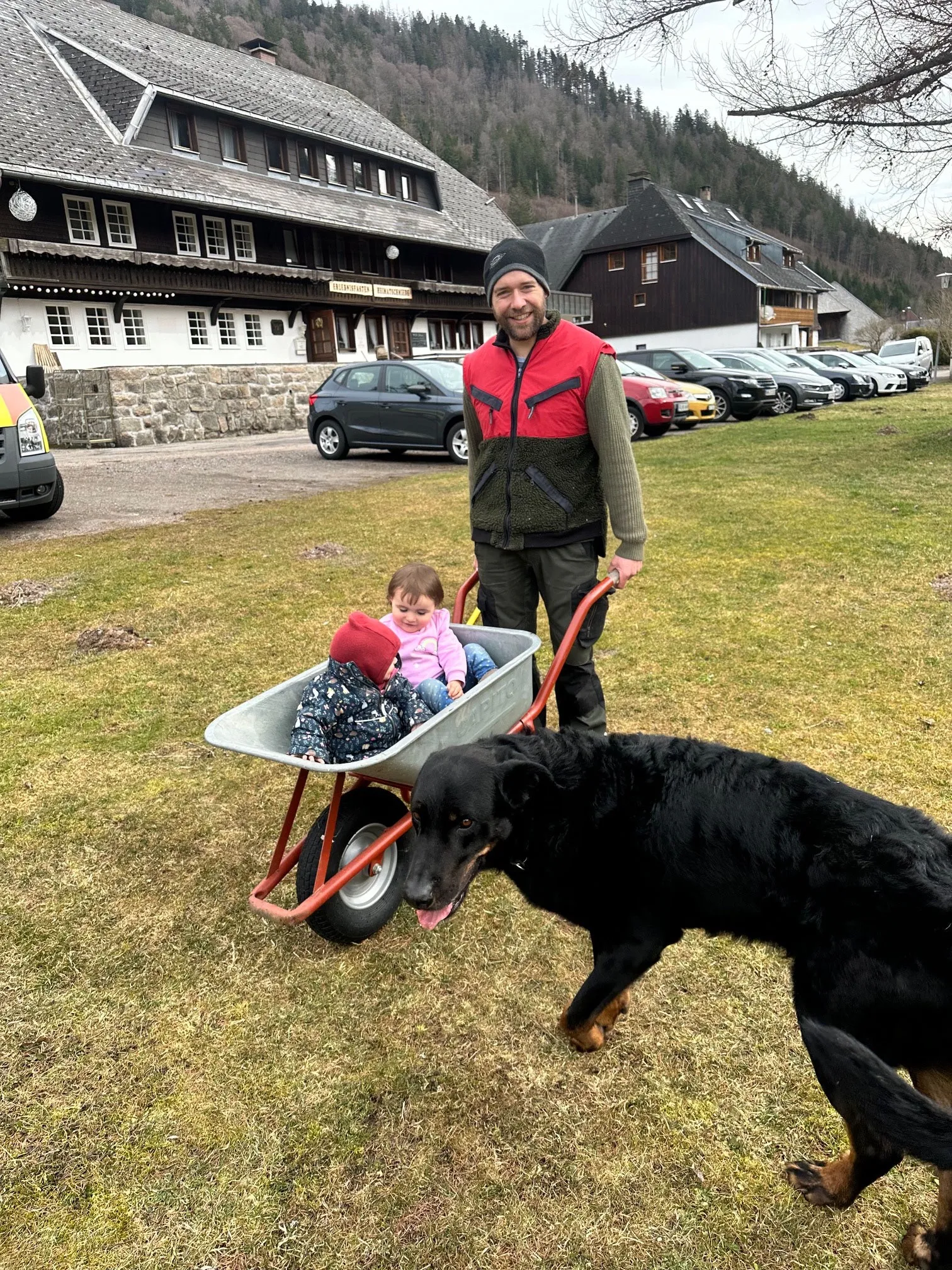 Paul schiebt eine Schubkarre über die Wiese vor dem Haus, neben ihm ein schwarzer Hund