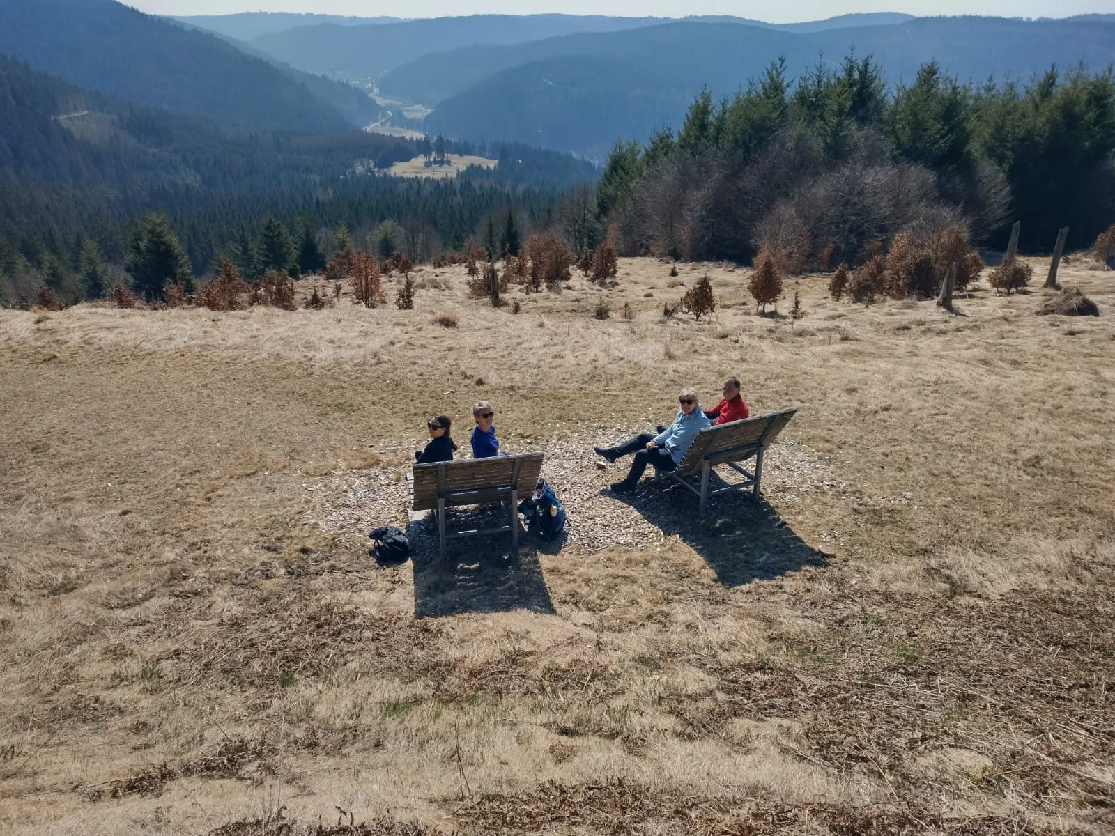 Vier Gäste sitzen auf zwei Holzbänken auf einer weiten, sonnigen Hochfläche und schauen hinunter ins Tal