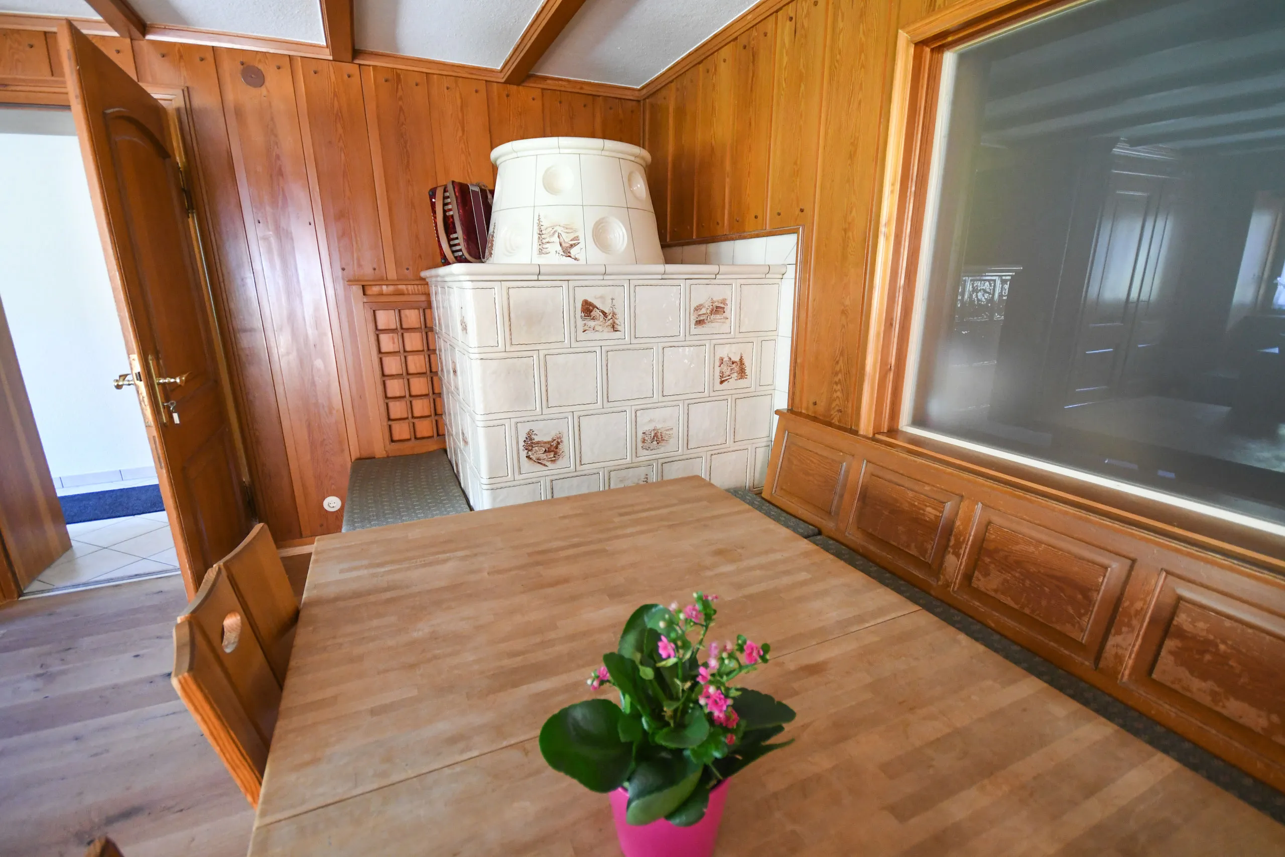 Die holzgetäfelte Stube mit weißem Kachelofen, einem großen Holztisch und Blumen