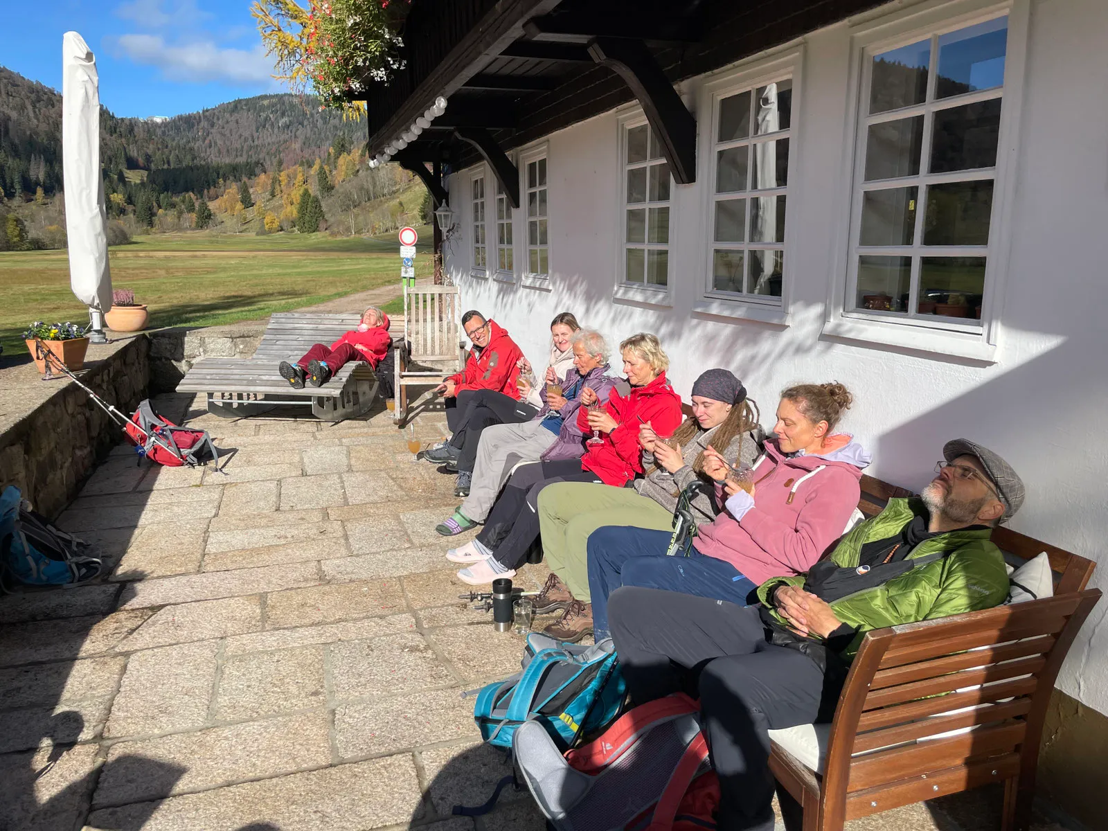 Gäste mit Rucksäcken sitzen in der Nachmittagssonne auf der Terrasse der Heimatschmiede, dahinter Wiesen und bewaldete Hänge